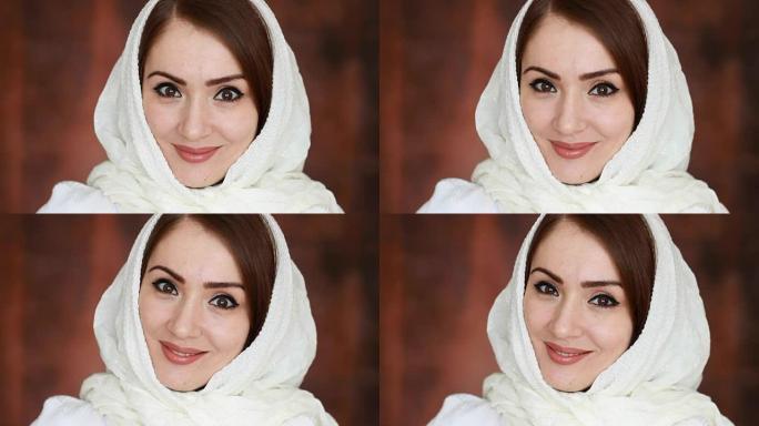 一位年轻而神秘的阿拉伯妇女的特写肖像穿着白色头巾，被称为头巾，微笑着看着相机。深棕色背景上美丽的穆斯