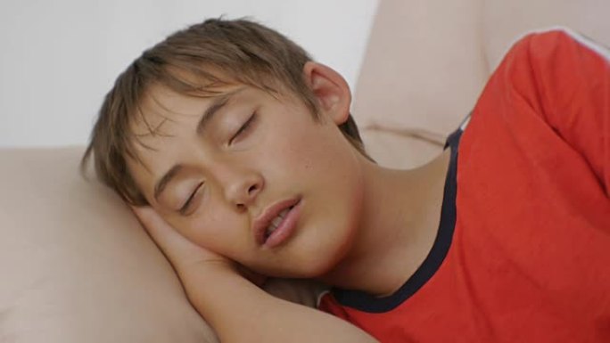 青少年睡在沙发上。白天穿着红色t恤的高加索少年男孩睡在米色真皮沙发上的特写。睡眠不足。