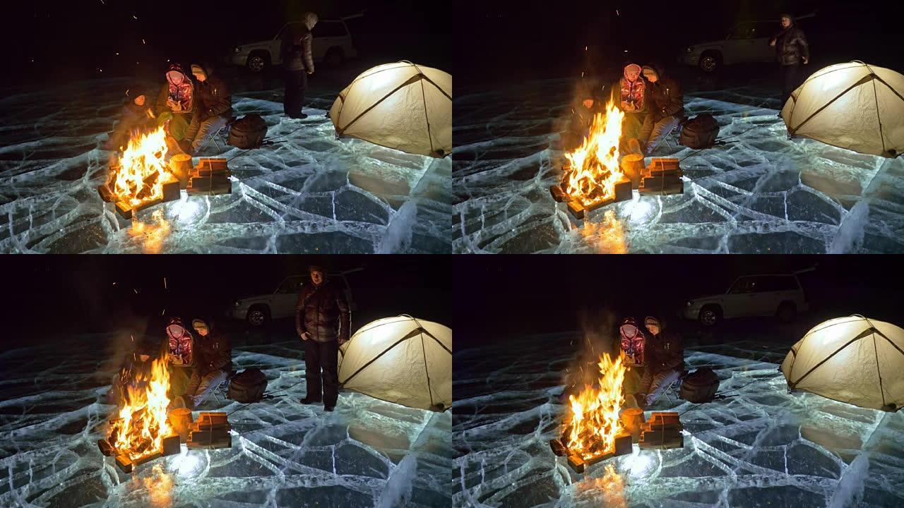 四个旅行者晚上在冰上着火。冰上的露营地。帐篷站在火旁边。人们在篝火旁变暖。摄影师在三脚架上拍摄。
