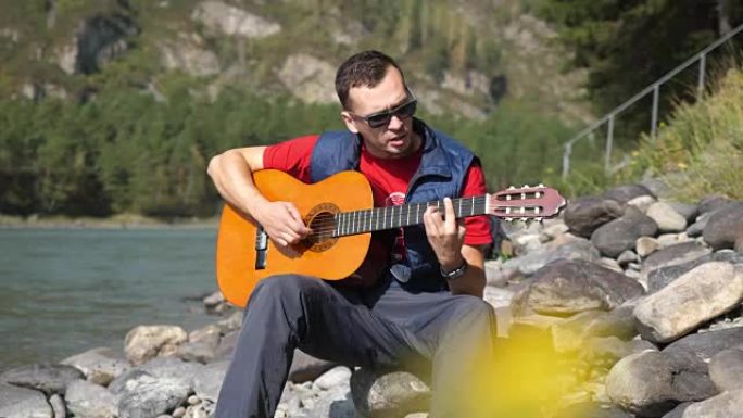 阳光明媚的日子，戴着墨镜的家伙坐在山河旁弹吉他。全高清，3840x2160
