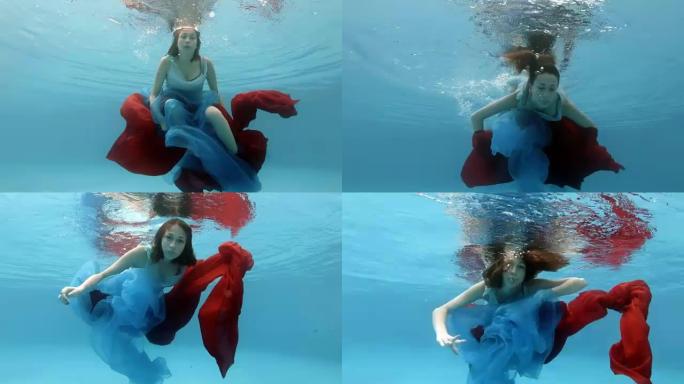 一个穿着连衣裙的美丽少女在游泳池的水下游泳，在水面附近用红蓝相布玩耍，看着相机。从下面看。动作摄像机
