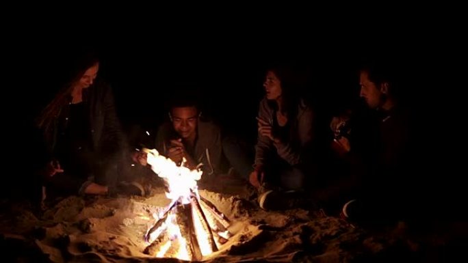 圆形摄像机运动: 多种族的年轻男孩和女孩在深夜坐在篝火旁唱歌和弹吉他。欢快的非洲裔美国人在炉火旁弹吉