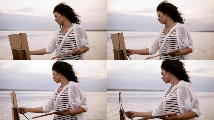 一个黑发卷发的美丽女人的特写镜头站在湖前，用调色板和画架画画。严肃而集中。早晨晴朗的天空