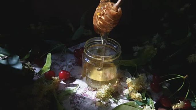 将蜂蜜倒入玻璃罐中，一束椴树花和木面上的红樱桃。一缕阳光。深色乡村风格。