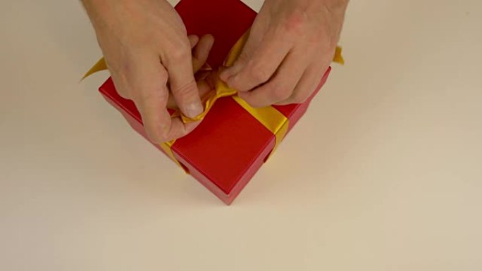 特写俯视图高角度。男士双手拉直礼品盒上缎带上的金蝴蝶结。为送礼活动做准备。带黄色蝴蝶结的礼品盒。红色