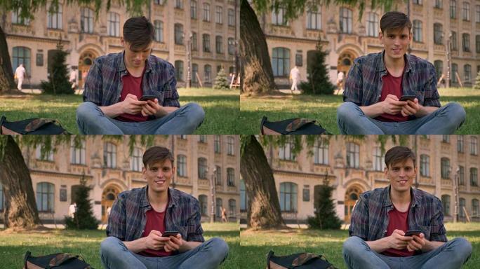 年轻英俊的男人坐在公园的草地上，点击智能手机，对着镜头，放松概念，交流概念，建立在背景上