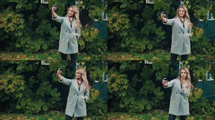 穿着灰色外套的金发女郎在秋季公园的智能手机上自拍