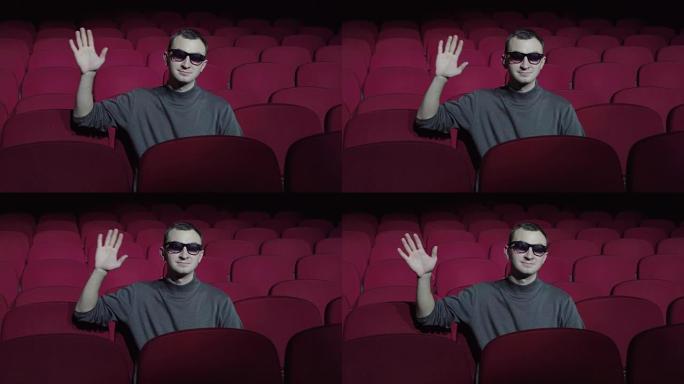 单身男子坐在黑暗电影院的舒适红色椅子上，向相机挥手
