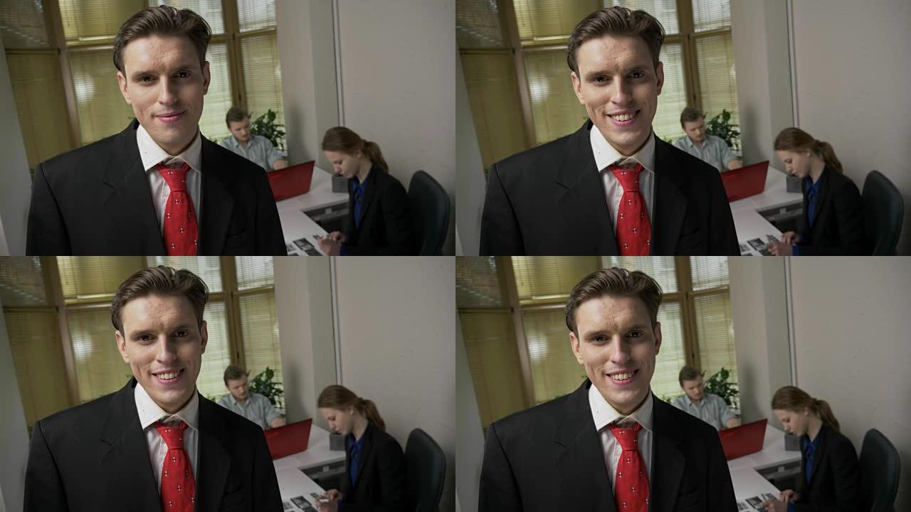 穿着西装的年轻人微笑着看着镜头，背景中的一个男人和一个女孩正在办公室里工作。60 fps