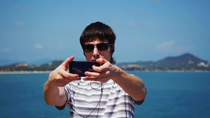 戴着墨镜的年轻人在海滩上用智能手机自拍，戴着耳机听音乐。苏梅岛。1920x1080