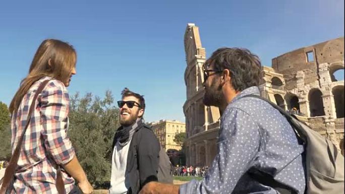 三个年轻的朋友游客坐在罗马斗兽场前，带着背包太阳镜在君士坦丁拱门附近玩得开心快乐美丽的女孩长发