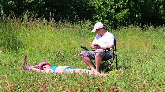一个穿着泳衣的女孩正在草坪上晒日光浴。在她旁边的野餐椅上，一个男人坐在智能手机上玩耍。拿一瓶水喝。