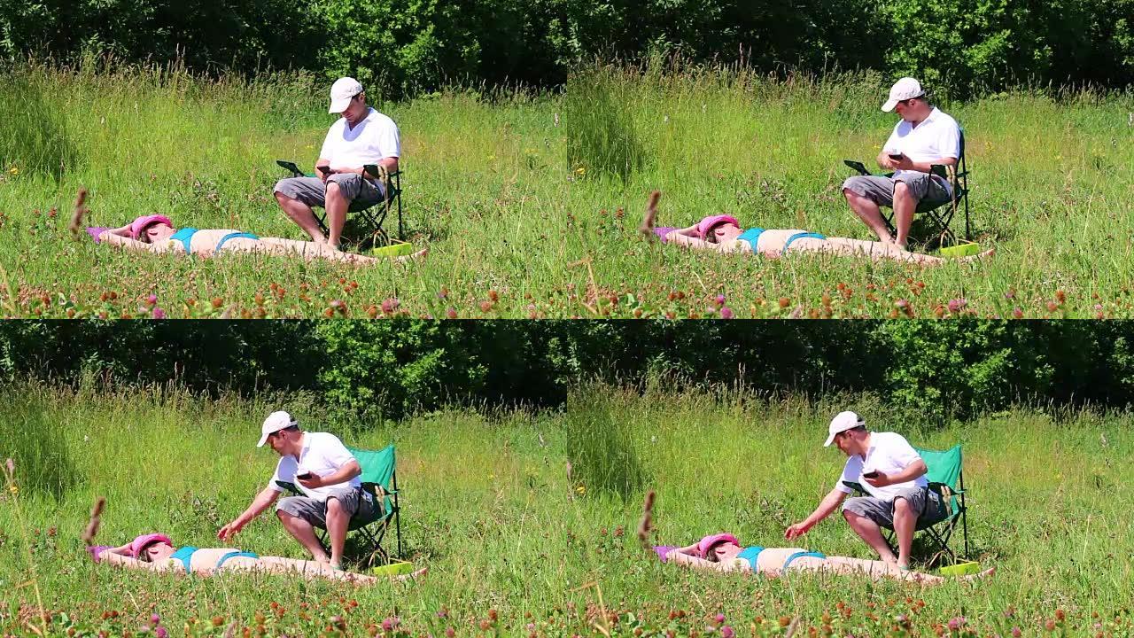 一个穿着泳衣的女孩正在草坪上晒日光浴。在她旁边的野餐椅上，一个男人坐在智能手机上玩耍。拿一瓶水喝。