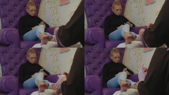 年轻漂亮的女人在水疗沙龙的修脚过程中使用智能手机