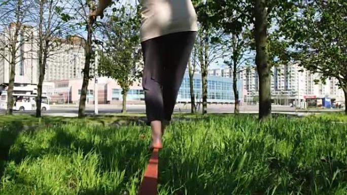 欧洲妇女正沿着城市公园的一条狭窄路线行走。女孩从后面平衡在松弛线上