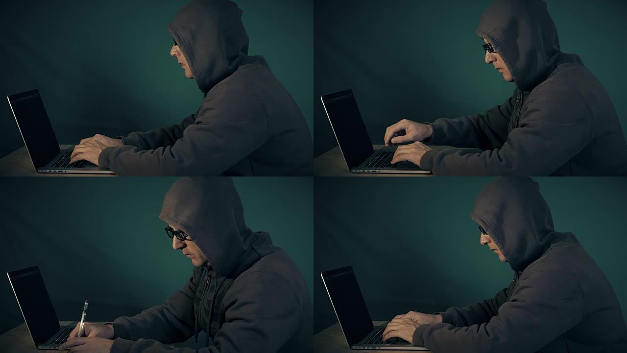 机罩和玻璃罩中的程序员在笔记本电脑的键盘上打字。黑客写备忘录。男人用手做了手势。低调的侧视图黑客坐在