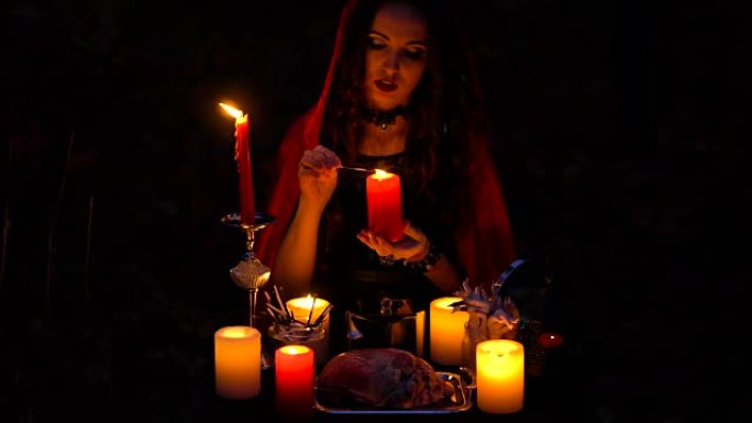 迷人的美丽黑发女巫，戴着红色斗篷的蜡烛，在神秘的秋天森林中让人联想到。带有心脏，鸡腿和豪猪针的巫术