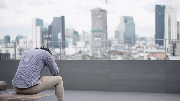 年轻的亚洲商人坐在办公楼屋顶露台的长凳上，对工作和生活问题感到压力和沮丧。背景中的城市视图。重性抑郁