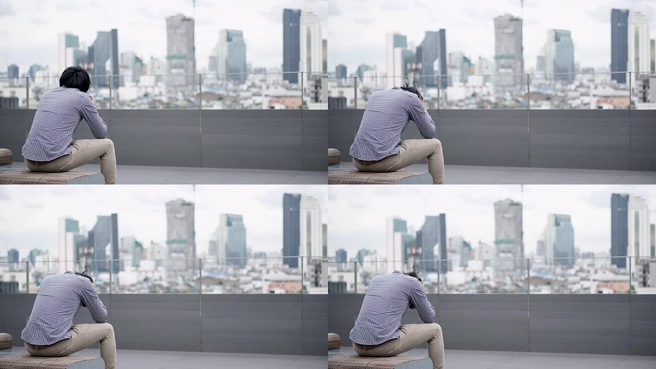 年轻的亚洲商人坐在办公楼屋顶露台的长凳上，对工作和生活问题感到压力和沮丧。背景中的城市视图。重性抑郁