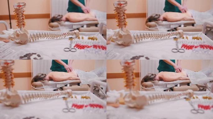 手动治疗师按摩一名躺在按摩台上的年轻女子，推动前景中的腰椎、脊柱模型和药物