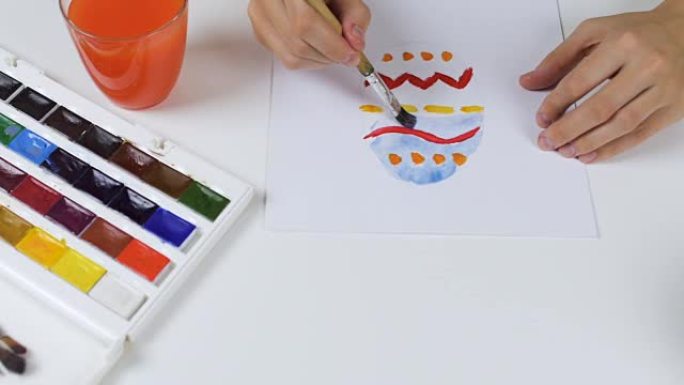 面目全非的女人在纸上画了一幅色彩鲜艳的素描，用于复活节卡片。桌子上是一个调色板，上面装有油漆和一杯水