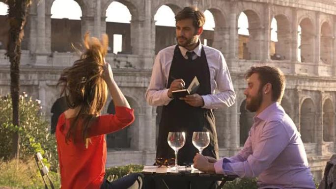 日落时分，浪漫优雅的情侣坐在罗马斗兽场前的餐厅餐桌旁，和服务员一起点菜和饮料