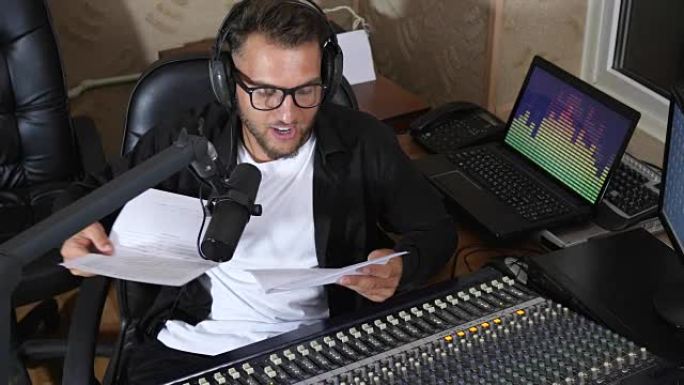 男性广播节目主持人用纸对着眼镜对着广播演播室设备附近的麦克风讲话