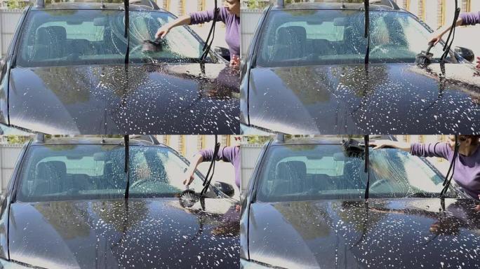 一个女人在晴天用刷子洗一辆黑色汽车。她把肥皂泡沫放在挡风玻璃上。