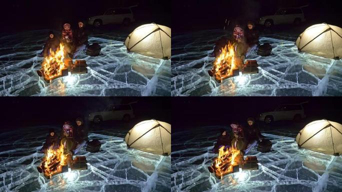 三个旅行者晚上在冰上着火。冰上的露营地。帐篷站在火旁边。贝加尔湖。附近有车。人们在篝火旁变暖，穿着睡