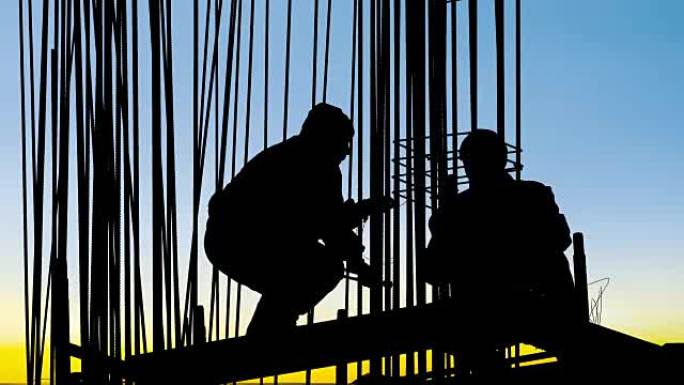 在金属框架后面，工人在日落时用电线将电线杆固定到建筑工地的钢筋混凝土支柱的底部