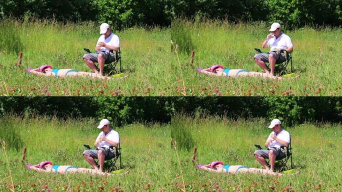 一个穿着泳衣的女孩正在草坪上晒日光浴。在她旁边的野餐椅上，一个男人坐着喝水。