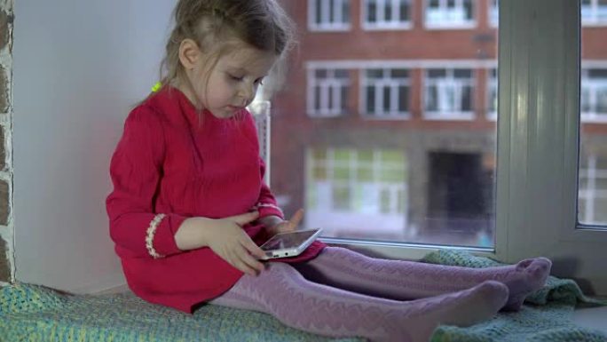 一个穿着粉色裙子的可爱小女孩坐在窗台上用智能手机