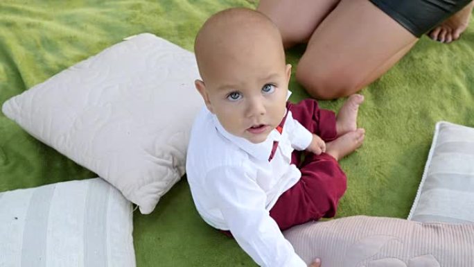 穿着漂亮大眼睛的婴儿看着相机坐在户外公园枕头附近的绿色格子上