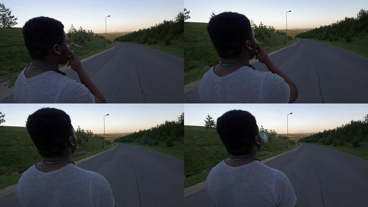年轻的非裔美国人在日落时走在空旷的道路上抽烟的后视图