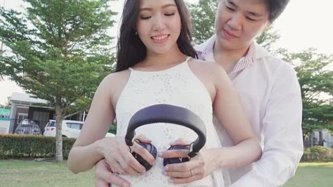 快乐的亚洲孕妇和丈夫在花园里的肚子上戴着耳机。4K