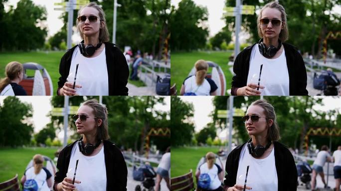 一个年轻漂亮的女孩穿过公园的前路画面。脖子上戴着耳机，戴着时尚的太阳镜。闲暇时间
