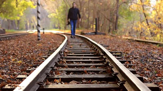 一个拿着DSLR相机的男人在背景的秋天森林中沿着火车轨道走。一名男子接近摄像机
