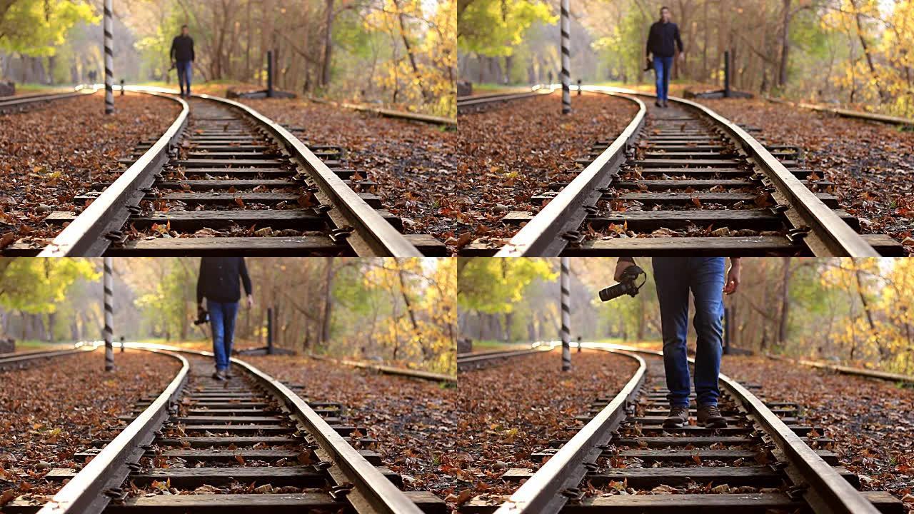 一个拿着DSLR相机的男人在背景的秋天森林中沿着火车轨道走。一名男子接近摄像机