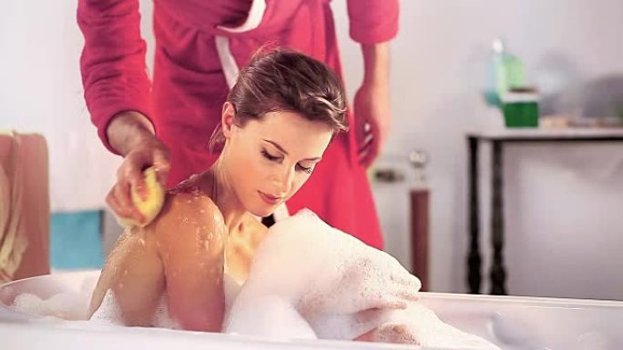 夫妇在浴室里放松，女人在浴缸里，男人用肥皂海绵摩擦肩膀