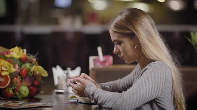 好女孩坐在咖啡馆的桌子旁，看着智能手机的屏幕，正在笔记本上做笔记
