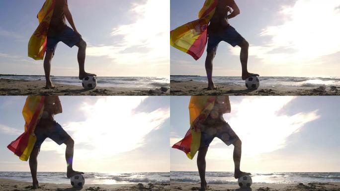 穿着西班牙西班牙国旗作为超级英雄斗篷的年轻人脚踩着足球站在海边的沙滩上，日落时看着海滩上的海洋摄像机