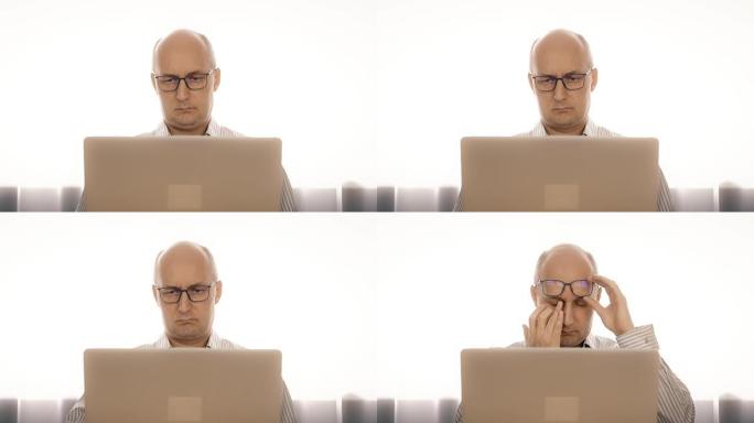 戴眼镜的悲伤男人在笔记本上工作，用乏力揉眼睛
