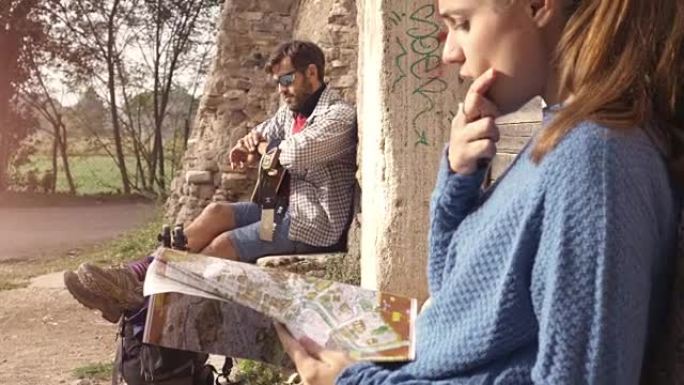 快乐的年轻夫妇背包客游客坐在罗马帕尔科德格里阿奎多蒂公园遗址中世纪建筑的木制门口附近，日出时带着吉他