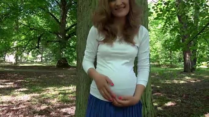 孕妇抚摸着她的肚子站在公园的大树干附近