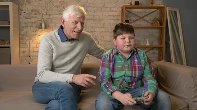 祖父看着孙子玩游戏机。老人和一个年轻的胖男孩坐在沙发上。电子游戏。家庭舒适，家庭田园诗，舒适概念，世
