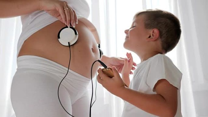 手拿手机的小男孩在灯火通明的房间里把耳机戴在孕妇的肚子上