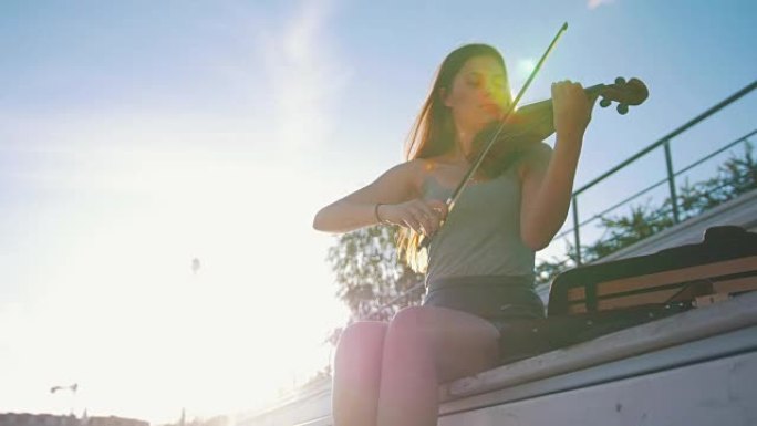 夏日公园里，年轻女子拉小提琴坐在长凳上，面对日落