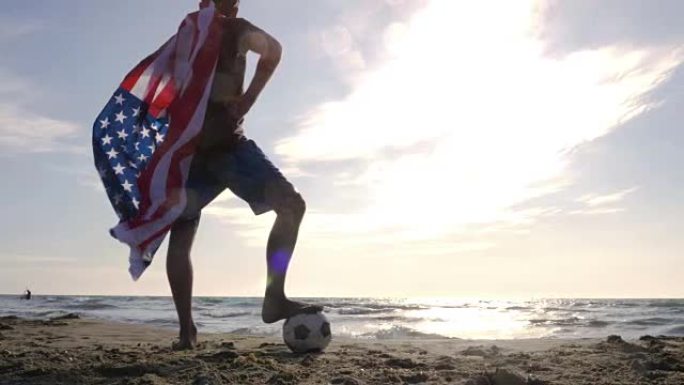 戴着美国美国国旗的年轻人作为超级英雄斗篷，脚踩着足球站在海边的沙滩上，日落时看着海滩上的海洋摄像机s