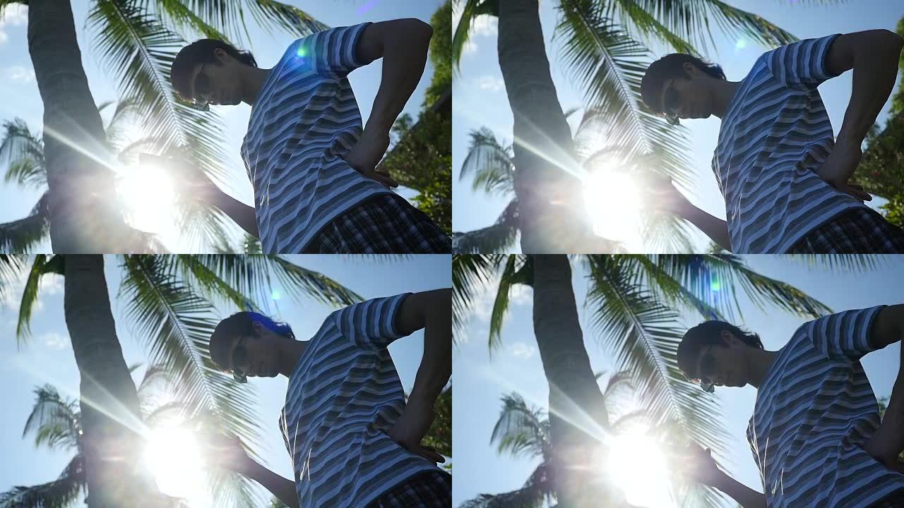 年轻人戴着墨镜，在室外的棕榈树下使用移动智能手机，具有阳光和镜头耀斑效果。慢动作。1920x1080
