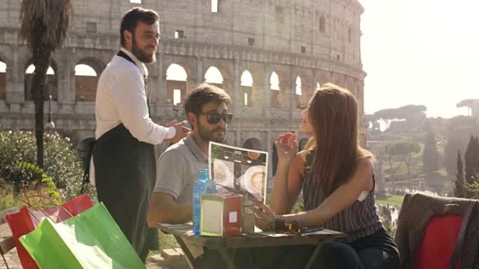 快乐的年轻夫妇游客在日落时坐在罗马斗兽场前的酒吧餐厅看菜单，叫服务员准备点食物和饮料